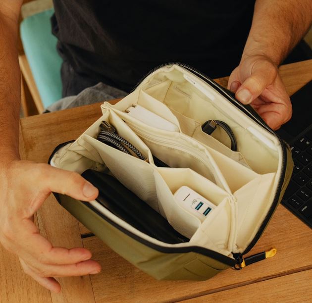 Blick in das weit geöffnete, helle Innenfach einer olivgrünen Peak Design Tech Pouch (Zubehörtasche in der Farbe Sage / Salbeigrün), die von einer Person auf einem hölzernen Tisch offengehalten wird. Die Lifestyle-Aufnahme verdeutlicht das strukturierte O