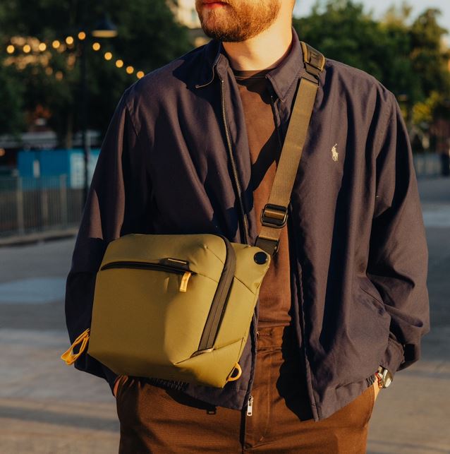 Eine Person im Halbprofil von vorne, die eine olivgrüne Peak Design Everyday Sling V2 Kameratasche (in der Farbe Kelp) cross-body an einem verstellbaren, dunklen Schultergurt trägt. Die wetterfeste Umhängetasche schmiegt sich eng an den Oberkörper an. Die
